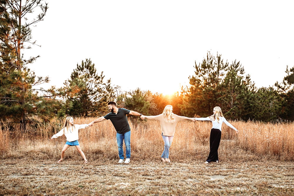 Family of four holding hands in golden field at sunset, professional photography with warm backlighting and pine trees