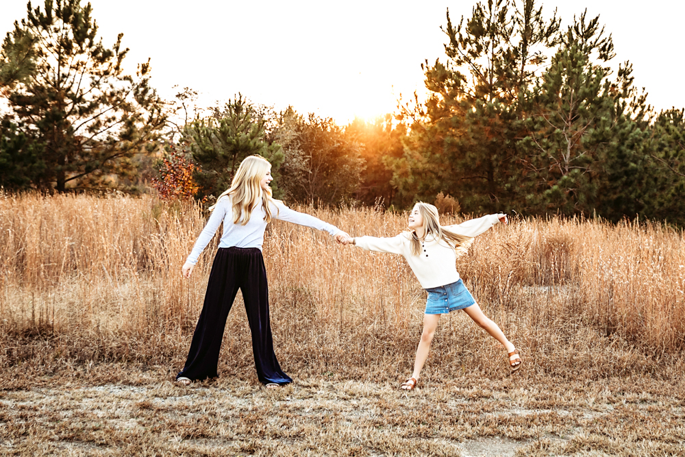 Adult and child holding hands in golden field at sunset, professional photographer captures warm family moment outdoors.