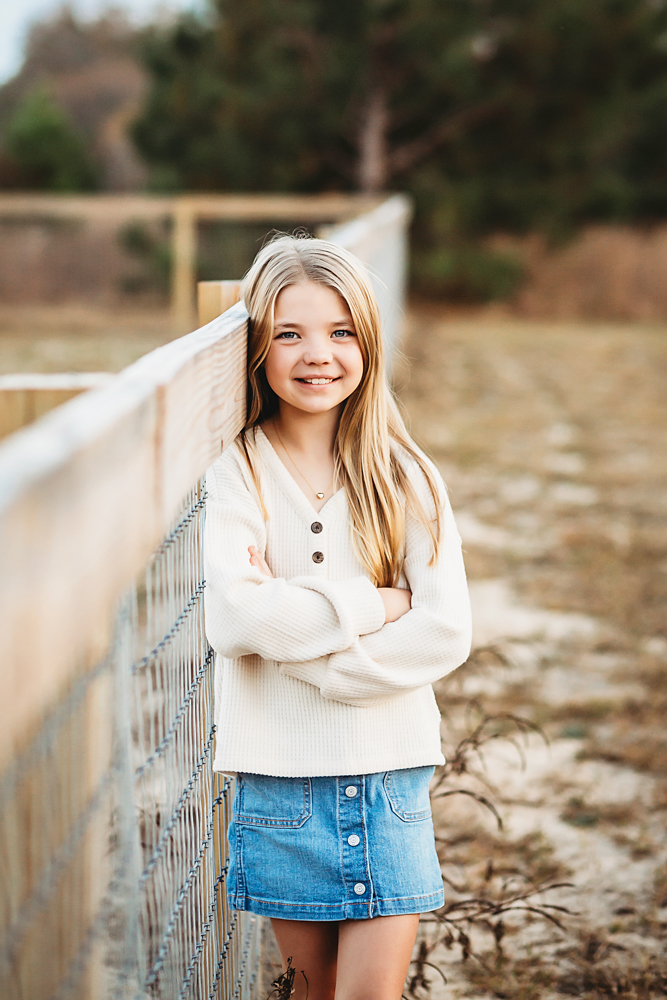 Smiling teen girl in cream sweater and denim skirt with arms crossed on wooden bridge during fall family photography