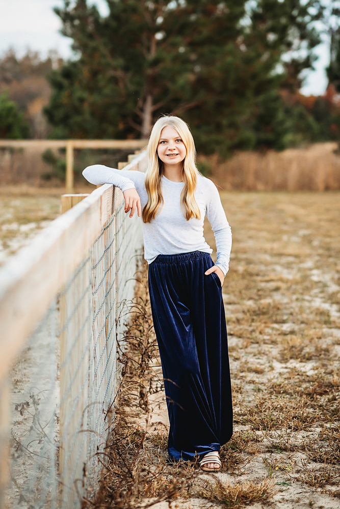 Smiling teen in white top and navy velvet skirt leaning on wooden fence in golden hour light - Reflection Images by Tracy Rowell