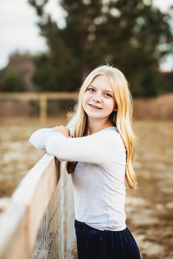 Smiling blonde teen in gray top leaning on wooden fence rail for fall family photos with soft natural lighting