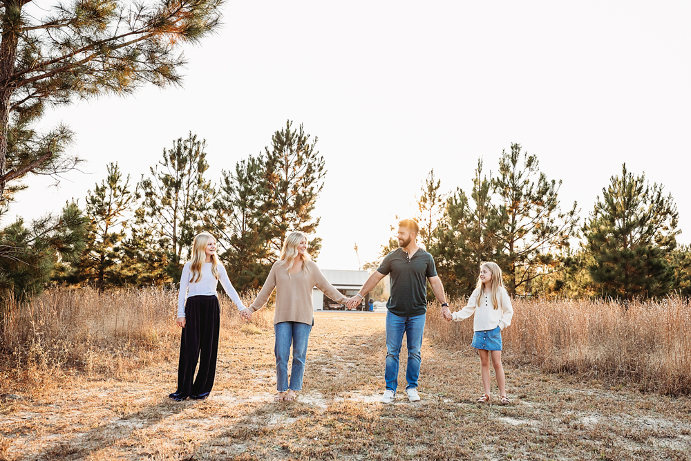 Professional fall family pictures: family of four holding hands in golden field with pine trees, warm sunset lighting