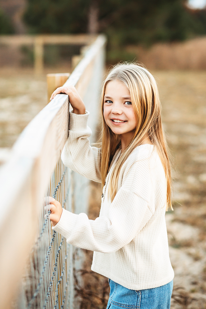 Smiling teen girl in cream sweater and jeans leaning on white fence, fall family pictures with warm golden lighting