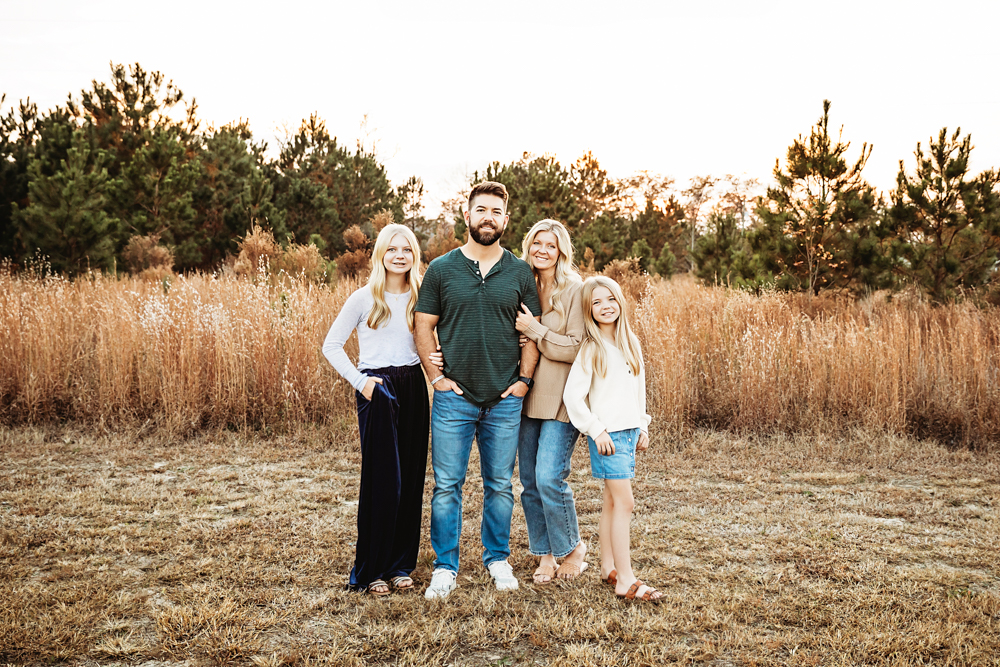 Family of four standing together in golden field at sunset, professional photography with warm lighting and autumn trees