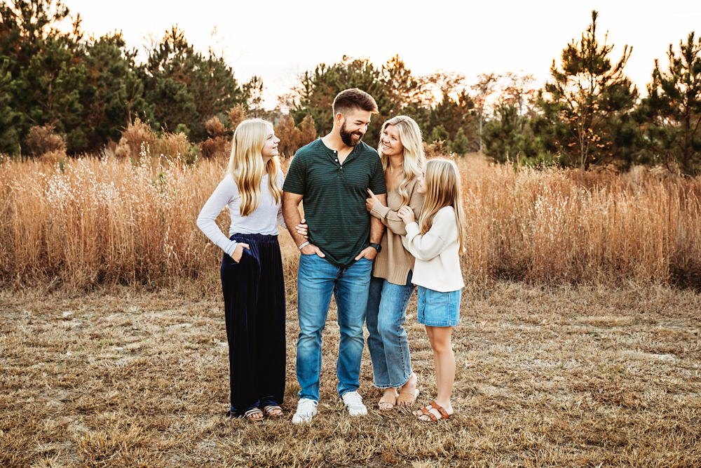 Family of four embracing in golden field at sunset, professional photographer captures warm moment with parents and teens