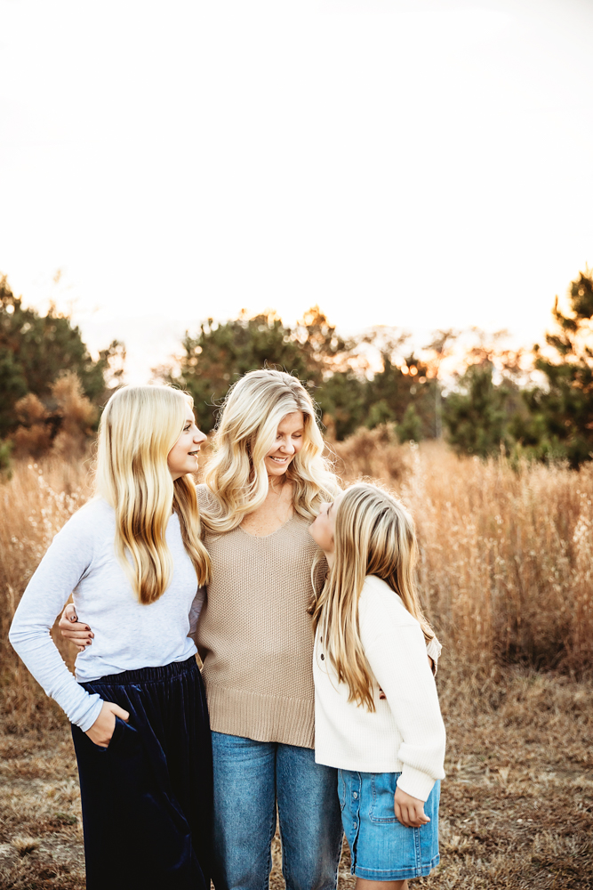 Professional fall family pictures of mother with two blonde daughters embracing in golden prairie field at sunset
