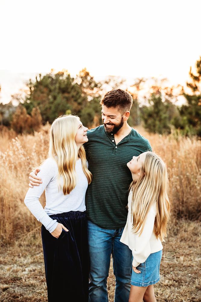 Fall family photography of smiling parents and child in golden field, wearing green henley and white tops with warm sunset lighting