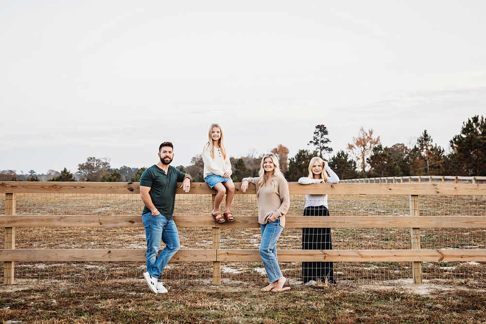 Family of four posing at wooden fence in rural setting, fall family photographer captures parents and teens in casual attire
