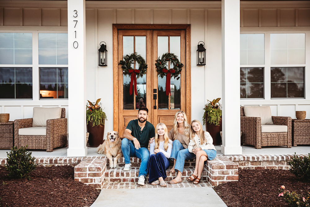 Fall family photos of four people and golden retriever sitting on front porch steps of white farmhouse with wreaths