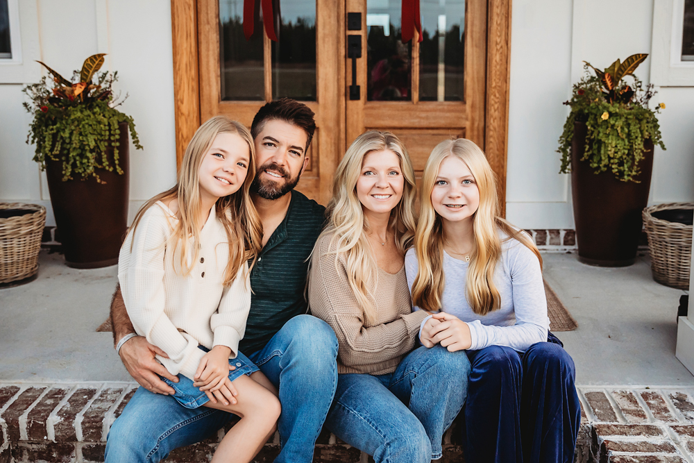 Fall family pictures: parents and two teens sitting on front porch steps in cozy sweaters and jeans with autumn decor