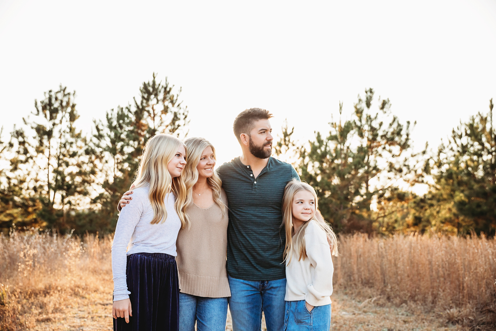 Fall family photography of parents with two teen daughters in golden field, warm evening light filtering through trees