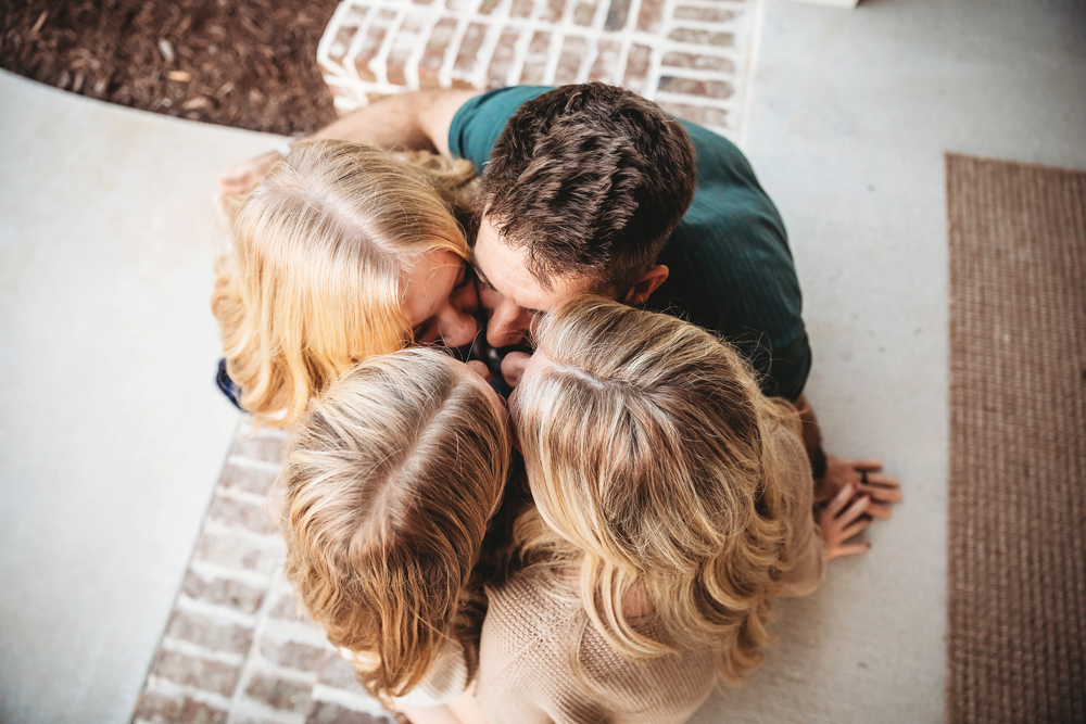 Family huddle on white bedding - parents and children with blonde hair embracing, warm lighting, professional photography near me