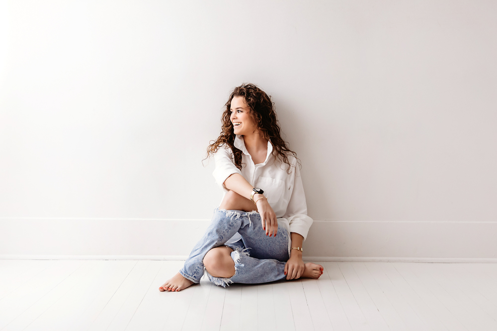 Young adult photography: smiling woman in white shirt and ripped jeans sitting cross-legged on white floor, soft studio lighting