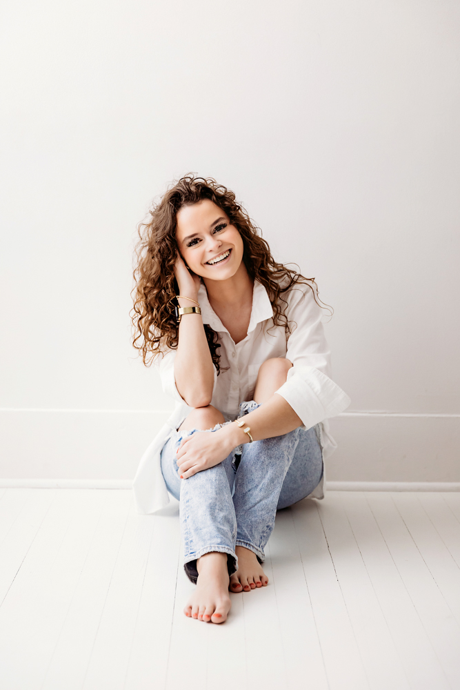Young adult photographer sitting cross-legged on white floor, curly brown hair, white shirt, blue jeans, bright studio lighting