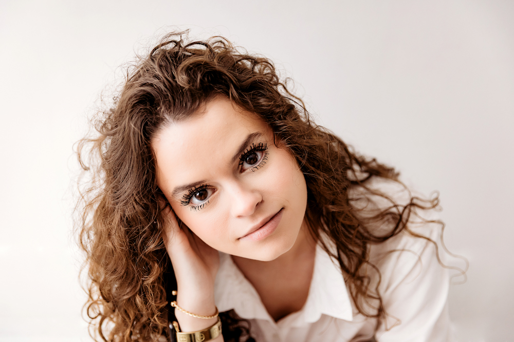 Young woman with curly brown hair resting chin on hand, wearing white top against soft gray backdrop - Reflection Images by Tracy Rowell