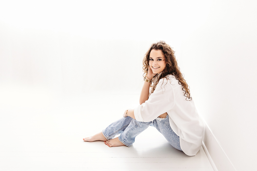 Teen girl in white oversized shirt and light jeans sitting cross-legged, smiling at camera on white backdrop - Tracy Rowell
