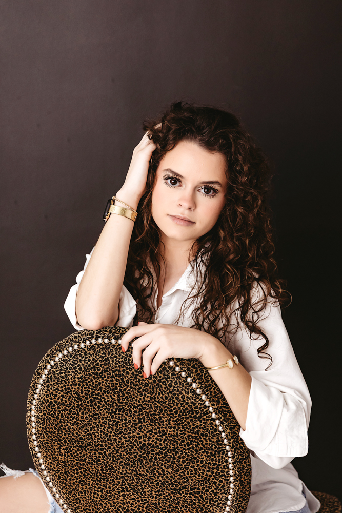 Teen with curly brown hair in white shirt posing with leopard print chair, moody lighting, florence sc newborn photographer