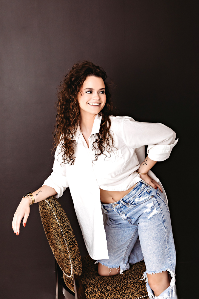 Smiling young woman with curly hair in white button-up shirt and ripped jeans, leaning on leopard chair, florence sc photographer