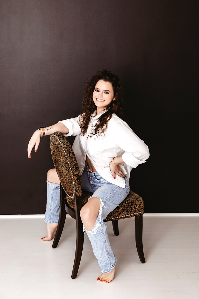 Smiling teen in white shirt and distressed jeans sitting on leopard chair, dark backdrop, professional photography studio near me