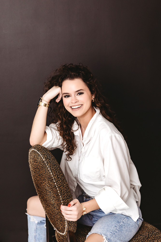 Smiling young woman in white shirt and leopard print pants sitting on stool, professional photography near me, warm studio lighting