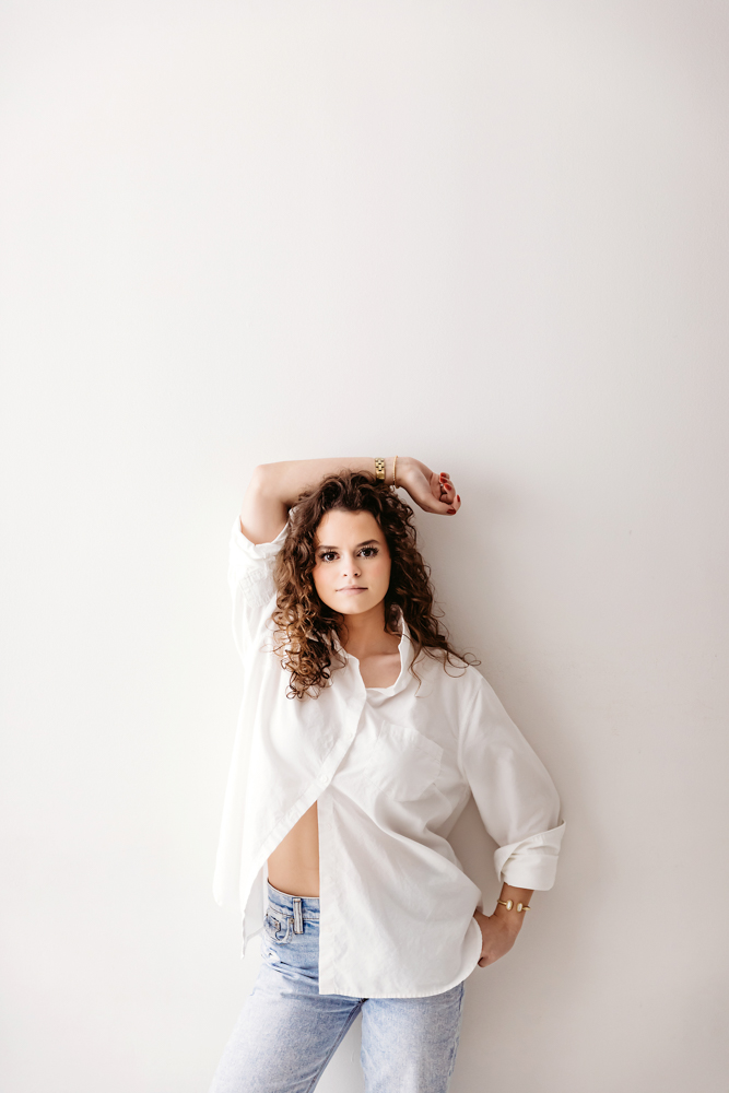 Teen with curly hair in oversized white shirt and jeans poses against cream backdrop, florence sc photographer portrait