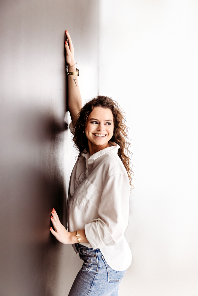 Smiling young woman in white hoodie and jeans poses against white wall, arm raised, curly hair flowing, professional photographer near me