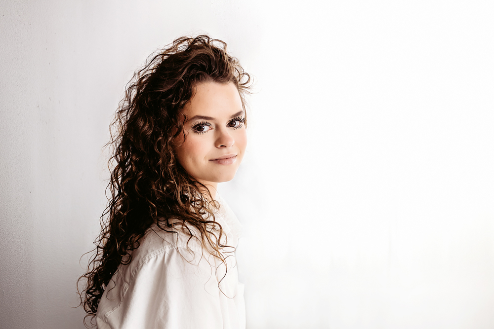 Young woman with curly brown hair in white shirt smiling at camera in professional photography studio with soft lighting
