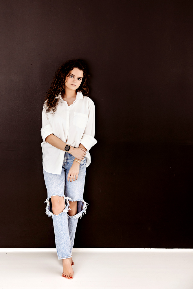 Professional photography of woman with curly hair in white shirt and distressed jeans against dark brown backdrop
