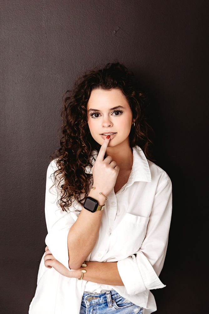 Young adult woman with curly hair in white shirt against dark wall, finger to lips pose by professional photographer