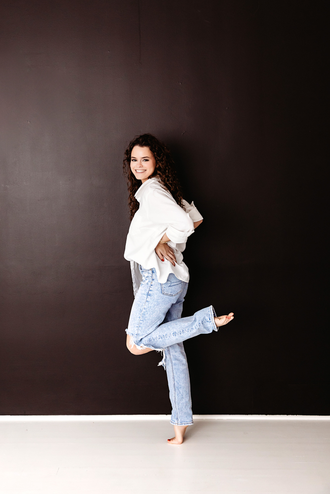 Smiling young adult with curly hair posing playfully in white shirt and jeans against dark wall - professional Young adult pictures