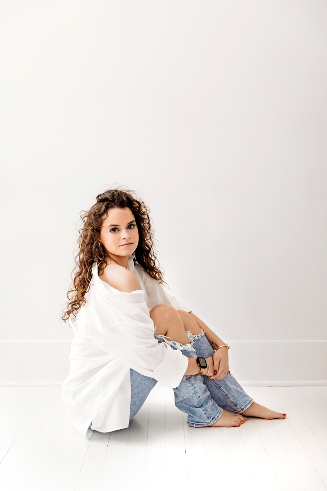 Young adult photographer portrait: woman with curly hair in white top and jeans sitting on white floor, soft lighting