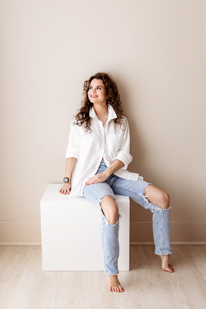 Adult woman with curly brown hair smiling while sitting on white cube, wearing white shirt and ripped jeans by Tracy Rowell
