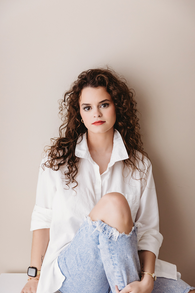 Young adult photos: Woman with curly brown hair in white button-down shirt and distressed jeans, seated against beige wall