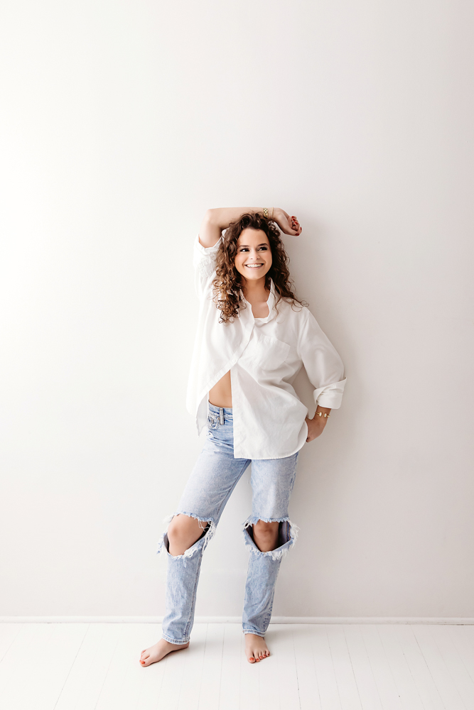 Smiling young woman in white button-up shirt and ripped jeans poses against neutral studio backdrop, professional photography studio near me