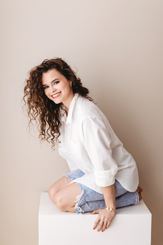 Young adult pictures: Smiling woman with curly brown hair in white shirt and distressed jeans sitting on cube, beige backdrop