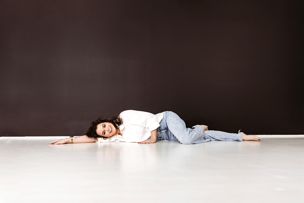 Smiling adult with curly hair lying on white studio floor in white top and jeans, professional photographer near me