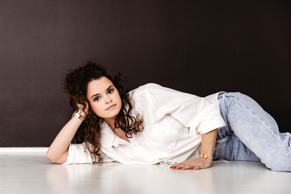Teen with curly hair lying on white floor in white shirt and jeans against dark backdrop in professional photography studio