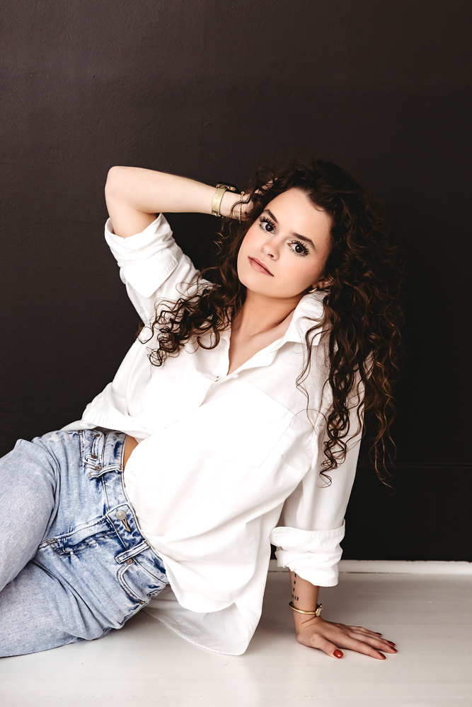 Young woman in white button-up shirt and jeans posed on floor against dark wall by professional photographer