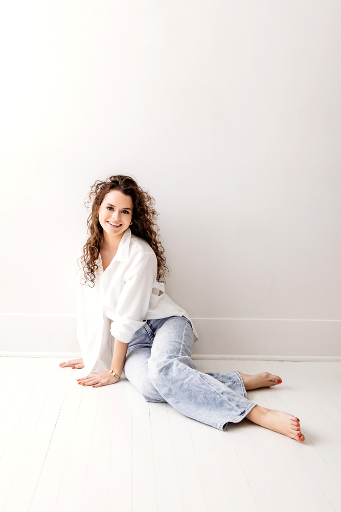 Professional young adult pictures: smiling woman in white shirt and light blue jeans sitting on white studio floor