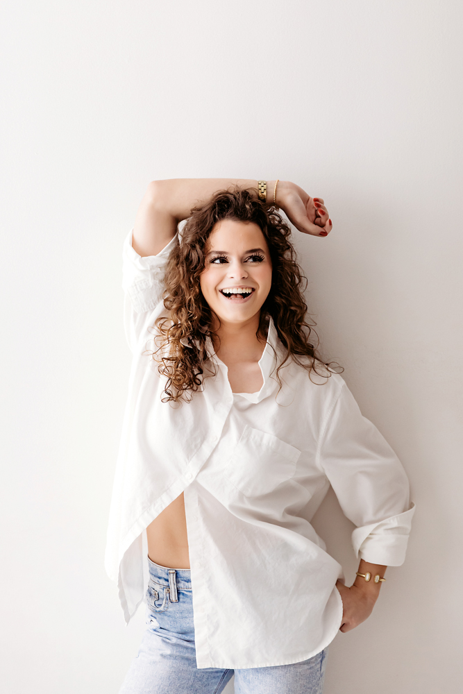 Laughing young woman with curly hair in white oversized shirt and jeans, hand in hair pose, soft lighting, professional photography near me