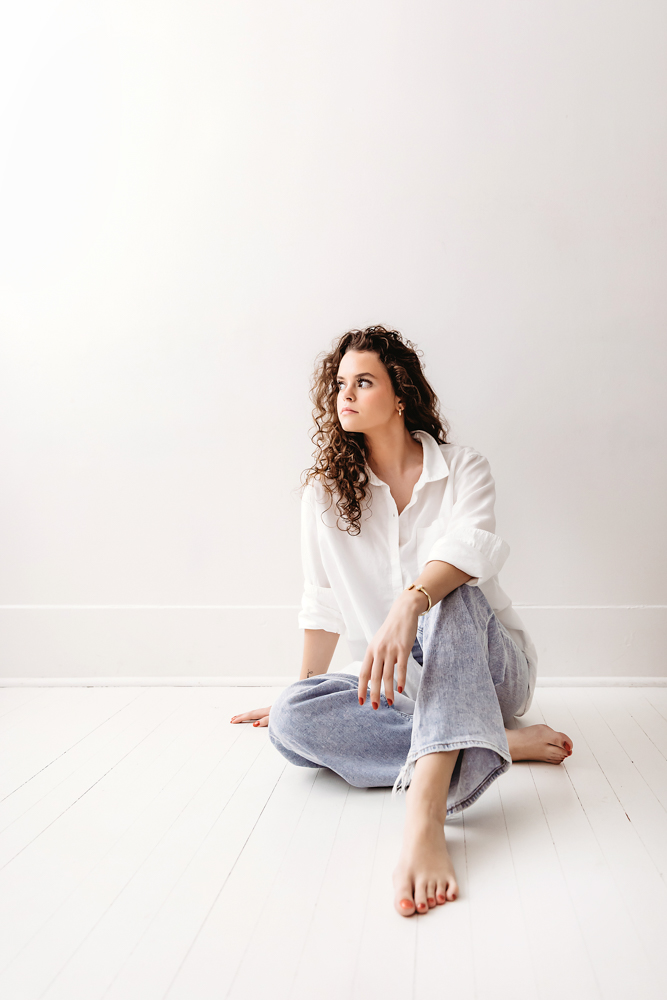 Young adult photography: woman with curly hair in white shirt and blue jeans sitting on white floor, soft studio lighting