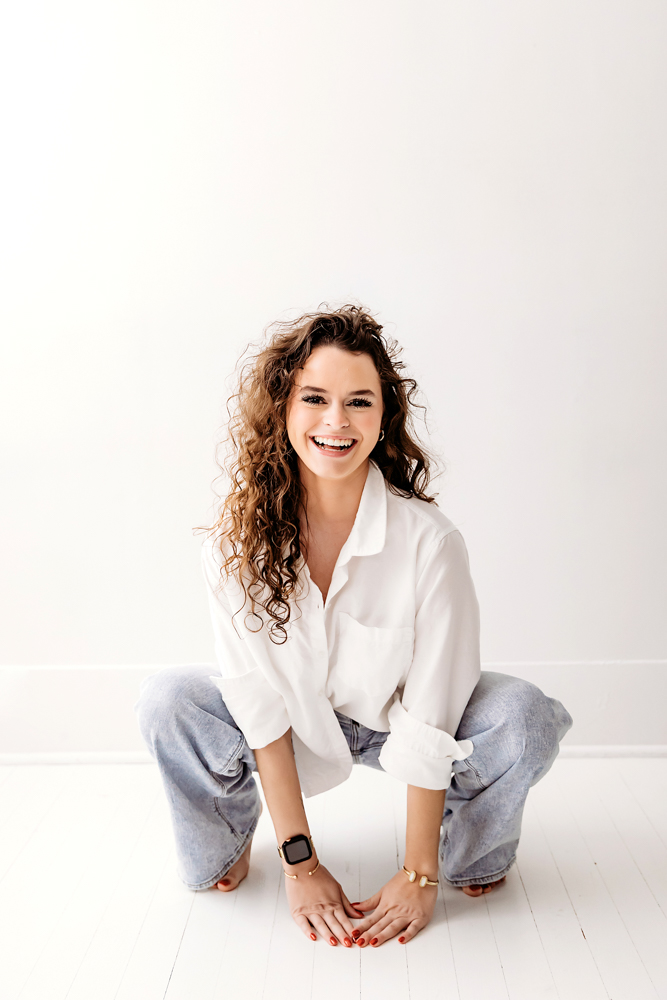 Young adult photographer with curly hair smiling, wearing white shirt and jeans, sitting cross-legged on white backdrop