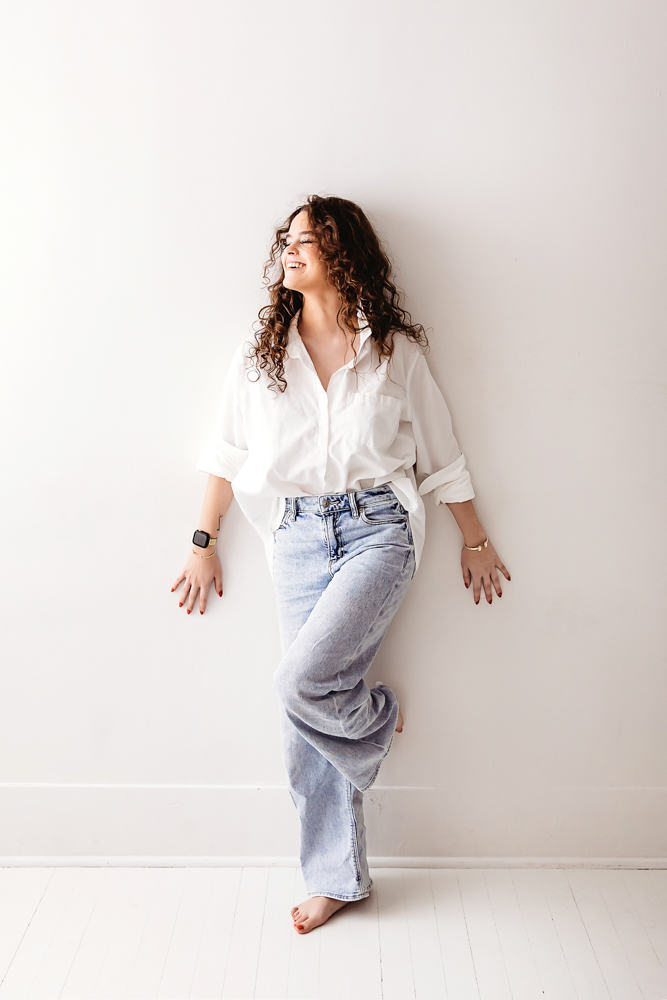 Woman with curly hair laughing against white wall, wearing white button-up shirt and light blue jeans, Tracy Rowell portrait