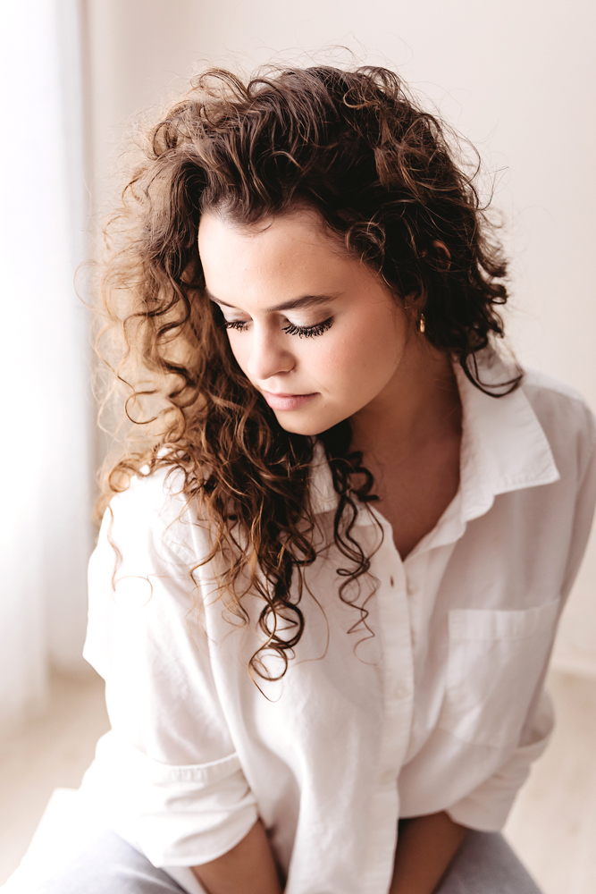 Young adult photos of woman with curly brown hair in white button-up shirt, eyes closed, soft natural lighting