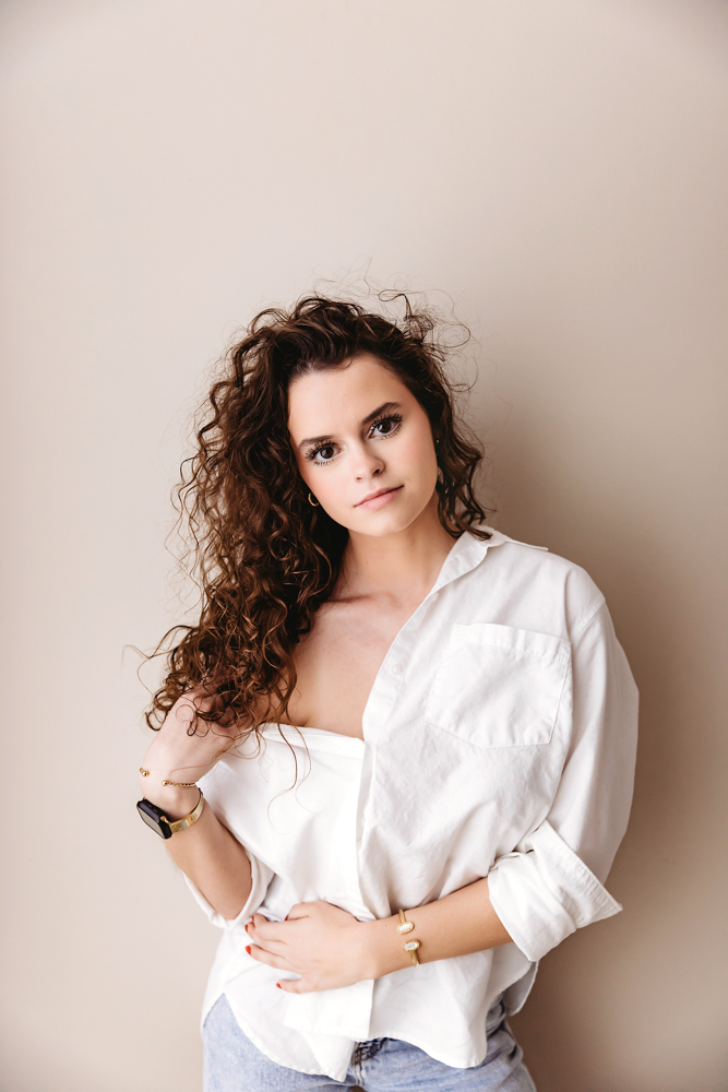 Young woman with curly brown hair in white button-up shirt poses against neutral backdrop by florence sc newborn photographer