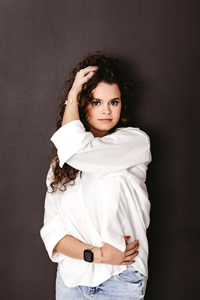 Young woman with curly brown hair in white oversized shirt and jeans posing against dark wall, professional photography studio near me