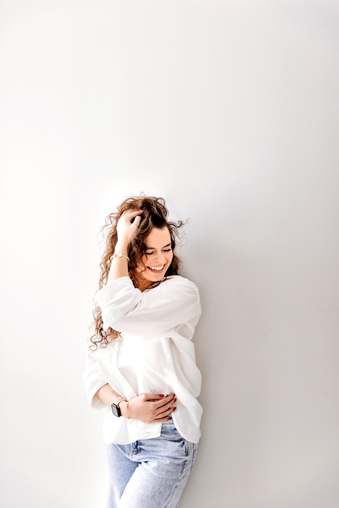 Smiling woman with curly brown hair in white sweater and jeans, hand in hair pose against light backdrop - professional photography near me