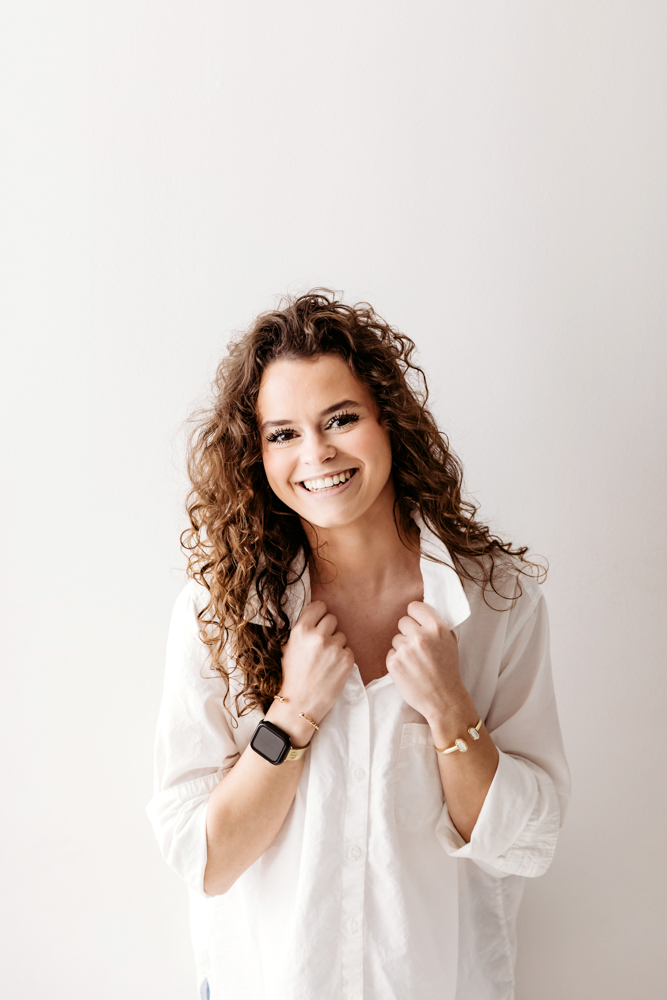 Smiling adult woman with curly brown hair in white button-up shirt on cream backdrop by professional photographer near me