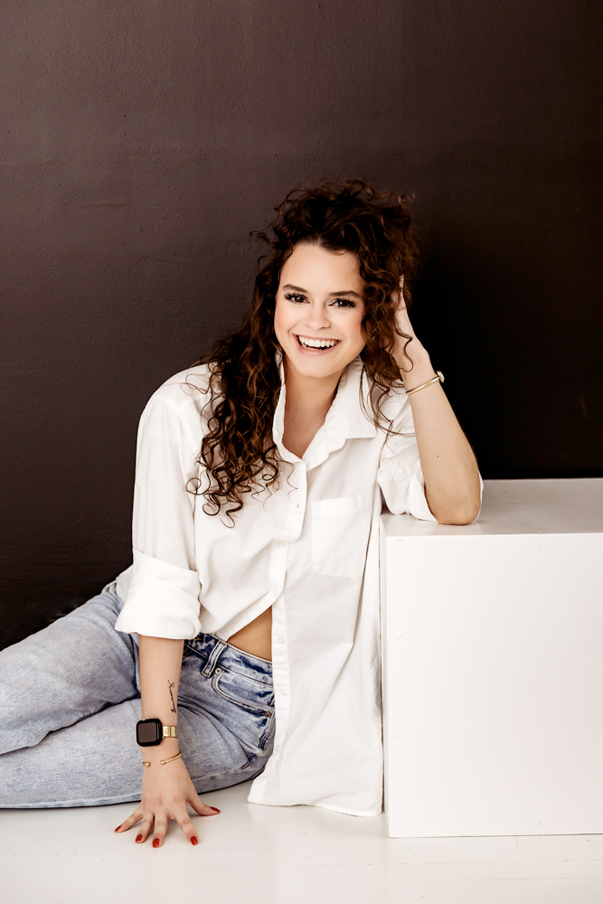 Smiling adult woman with curly brown hair in white button-up shirt and jeans, sitting on white surface against brown wall by professional photographer near me