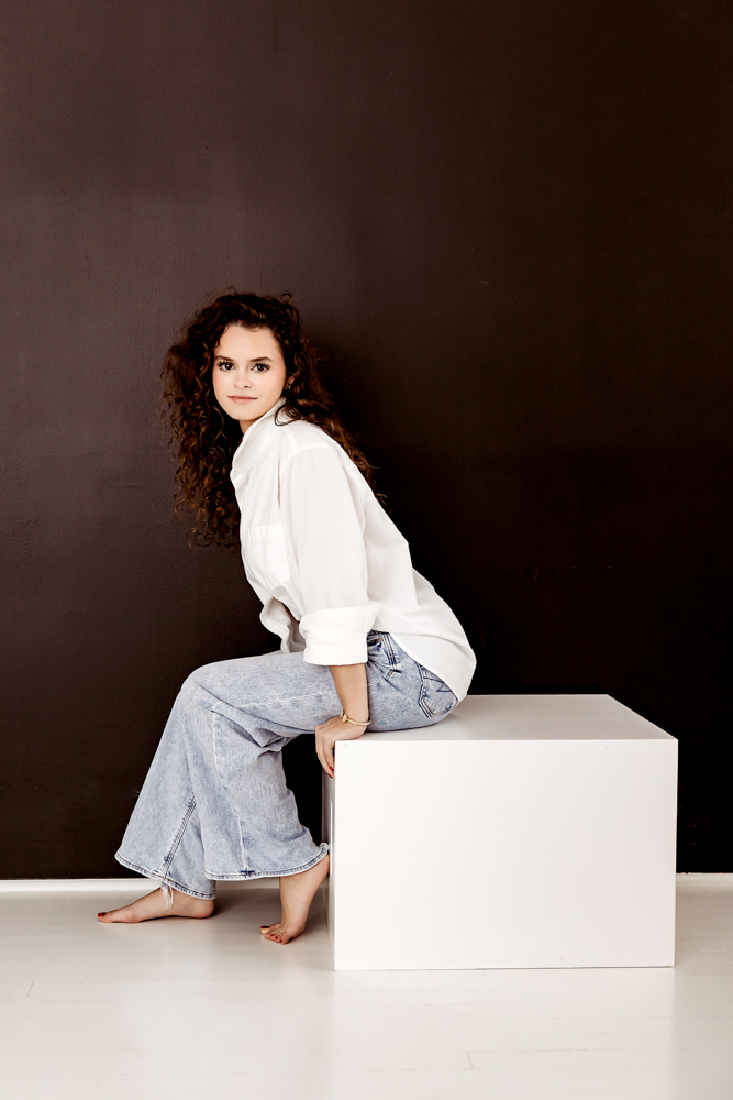 Teen with curly hair in white shirt and light jeans sitting on white cube against dark backdrop in professional photography studio
